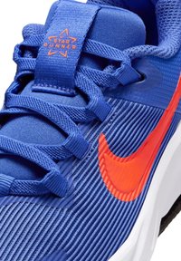 Sininen urheilukenkä, jossa on teksturoitu yläosa, kirkkaan oranssi Nike-logolla varustettu swoosh, vahvistettu varvasosa ja tähti-logo kielen päällä.