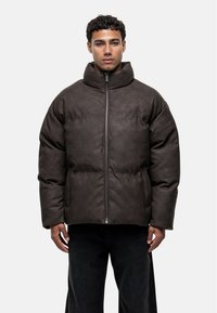 Veste rembourrée marron avec un col montant, fermeture à glissière sur le devant et deux poches latérales. Fabriquée en matériau lisse et texturé avec des détails de couture.