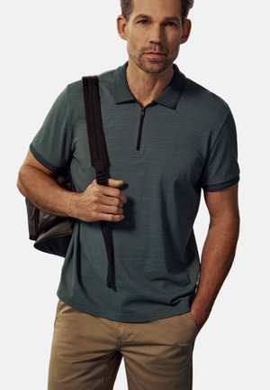 Homme portant un polo vert foncé texturé et un pantalon beige, avec un sac à dos noir porté sur une épaule, au visage neutre.