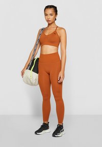Reggiseno sportivo marrone e leggings a vita alta realizzati in tessuto elastico. Porta una borsa beige e nera con un accento verde. Scarpe sportive nere.