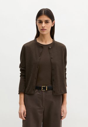 Vrouw draagt een donkerbruine vest over een bijpassende top en broek, met een zwarte riem en goudkleurige gesp, staand tegen een effen witte achtergrond.