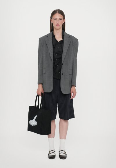 Over-sized grå stribet blazer over en sort skjorte med knapper, parret med navy-stribede shorts, hvide sokker og en sort tote-bag med grafik.