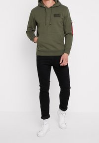 Hoodie verde oliva con tasca frontale, cappuccio regolabile con cordino e tasca con zip sulla manica con etichetta rossa. Jeans neri slim-fit e sneakers bianche.