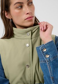 Grüne Jacke mit Kragen und einem Ärmeldetail aus Denim. Verfügt über silberne Beschläge und eine glatte Textur. Nahaufnahme zeigt Knopfdetails und Kragenstil.