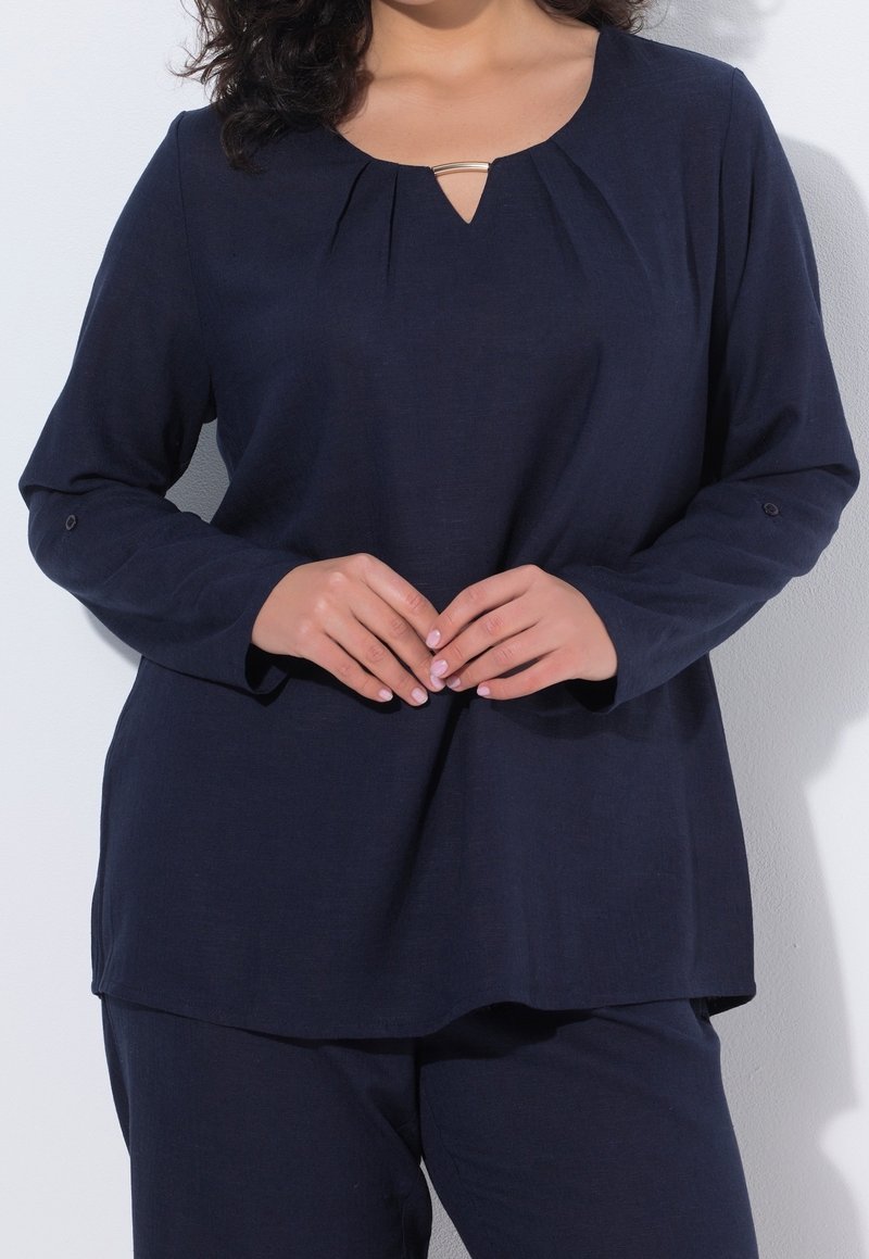Femme portant un chemisier bleu marine ample à manches longues avec une petite encolure goutte d'eau et un pantalon bleu marine assorti, les mains jointes à la taille.