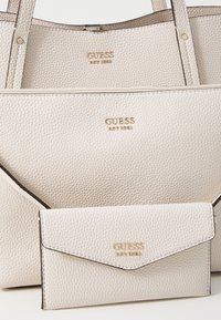 Tři bílé texturované kožené tašky a peněženky Guess s pozlaceným logem uspořádané do sebe.