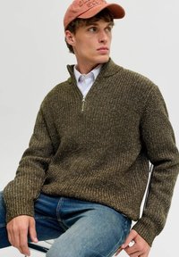 Junger Mann in olivgrünem Strickpullover mit halb-zipp, blauen Jeans, weißem gestreiftem Hemd und ausgebleichtem roten Cap, sitzt auf einem Stuhl und schaut nach rechts.