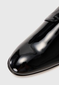 Zapato de dress de charol negro brillante con punta afilada y detalles de cordones sobre un fondo gris claro.