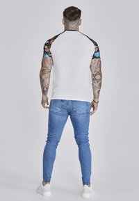 White fitted t-shirt with black floral pattern sleeves, paired with blue skinny jeans and white sneakers. Smooth texture, casual design.