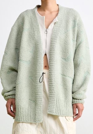 Person iført en oversized lysegrøn struktureret cardigan med åben front over en hvid lynet top og lyse beige bukser.