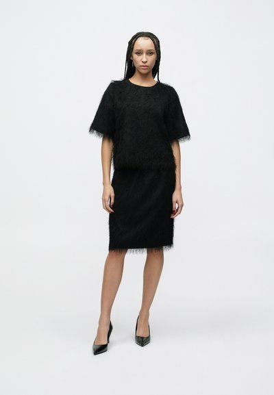 Tenue noire avec texture duveteuse, haut à manches courtes et jupe longueur genou. Associée à des escarpins noirs pointus. Design simple et épuré.