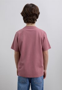 Różowy bawełniany t-shirt z okrągłym dekoltem i krótkimi rękawami, pokazany od tyłu na dziecku, zestawiony z niebieskimi jeansami.