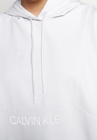 Haut à capuche blanc avec cordons et logo texte "CALVIN KLEIN" sur la poitrine, porté par une personne vue de la mâchoire au torse.