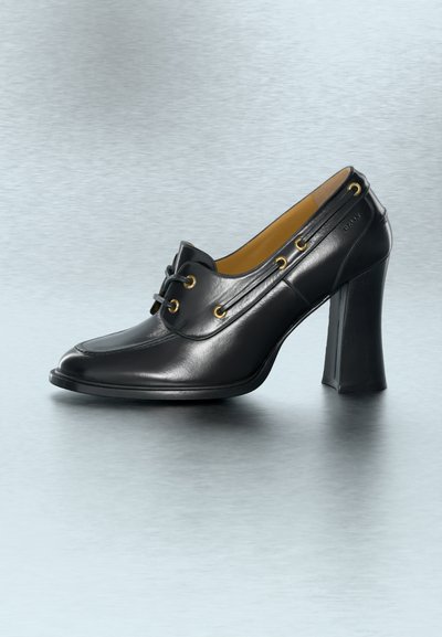 Μαύρο δερμάτινο γυναικείο παπούτσι με ψηλό τακούνι, χρυσές τρύπες για κορδόνια, μπροστινό δέσιμο με κορδόνια, στρογγυλεμένη τετράγωνη μύτη και ευρύ μπλοκ τακούνι σε γκρι φόντο.
