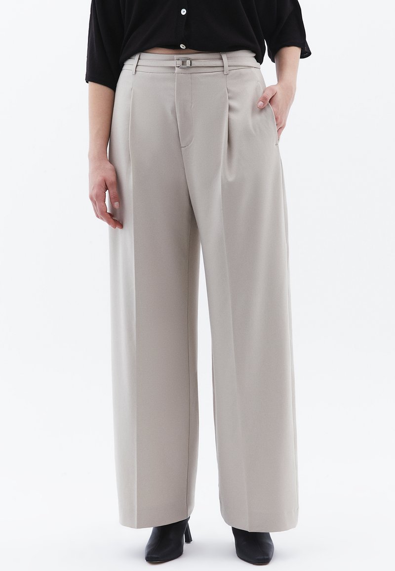 Pantaloni beige a gamba larga con vita alta, pieghe frontali e un piccolo dettaglio in argento sulla cintura. Tessuto morbido, linee pulite.