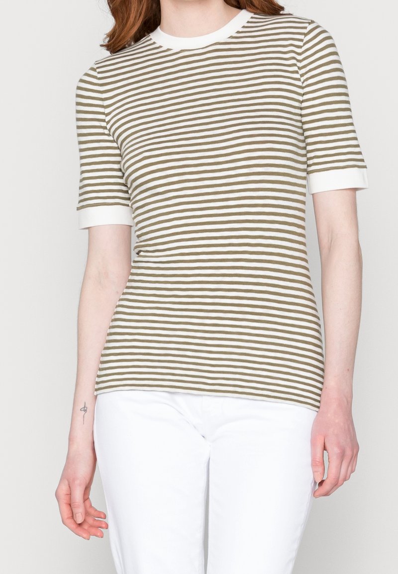 Mujer vistiendo una camiseta de manga corta a rayas en verde oliva y blanco, con puños y cuello blancos, combinada con pantalones blancos, de pie frente a un fondo liso.