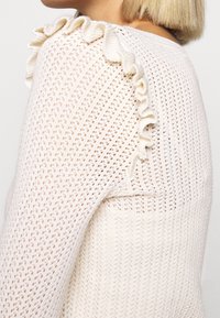Žena v krémovém pleteném svetru s krajkovým vzorem a volány podél švu na rameni, zobrazená z boku a zezadu.