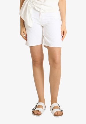 Donna che indossa pantaloncini bianchi testurizzati al ginocchio, top bianco arricciato e sandali bianchi con doppia fibbia, in piedi su uno sfondo semplice.