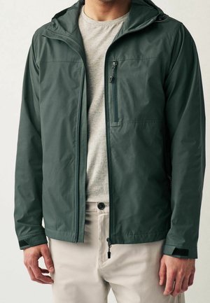 Mężczyzna ubrany w ciemnozieloną kurtkę z kapturem z zamkami na piersi i na przodzie, na jasnoszarej koszulce i beżowych spodniach.