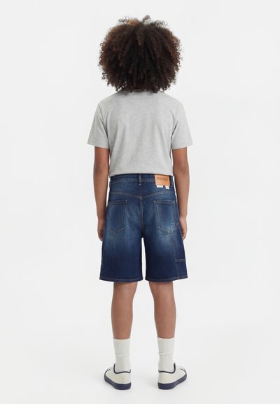 Παιδί με σγουρά μαλλιά στέκεται με την πλάτη, φοράει γκρι μπλουζάκι, σκούρα μπλε τζιν σορτς, λευκές κάλτσες και λευκά αθλητικά με ναυτικό περίγραμμα.