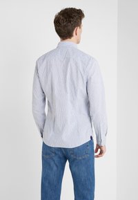 Hombre vestido con una camisa de manga larga a rayas en azul claro y blanco, y jeans azules, de pie frente a una pared blanca con la espalda hacia la cámara.