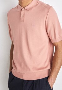 Uomo che indossa una polo a maglia leggera rosa chiaro a maniche corte con bottoni e logo "BOSS" sul petto, abbinata a pantaloni scuri.