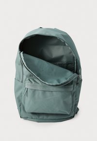 Mochila de tecido verde com um design simples, apresentando um fecho superior com zip, um bolso frontal e um interior espaçoso para armazenamento.