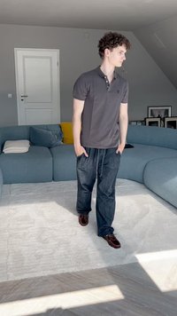 Graues Poloshirt, dunkle Jeanshose und braune Schuhe. Im Hintergrund sieht man ein hellblaues, geschwungenes Sofa und einen hellen Teppich auf einem Holzboden.