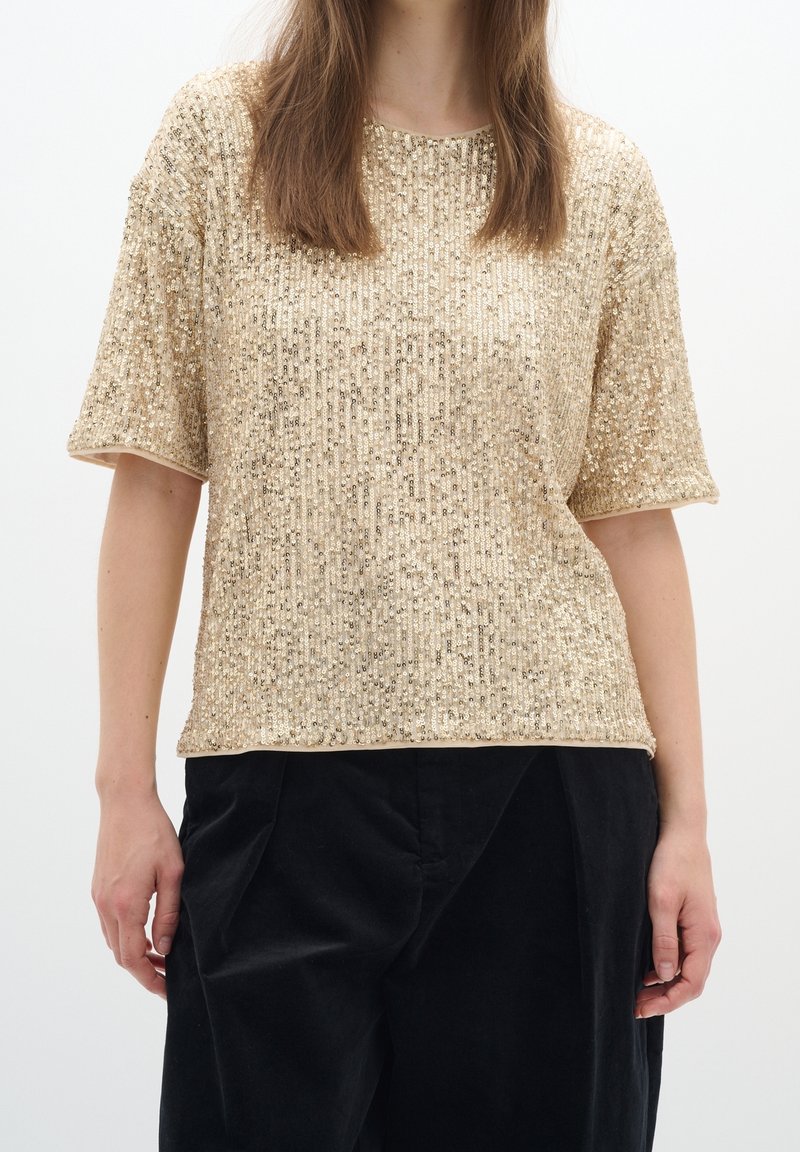 Femme portant un haut à manches courtes en sequins dorés associé à un pantalon noir ample, vue du cou aux hanches sur fond blanc.