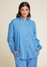 Lys blå denimskjorte med knapper foran, lange ermer og klassisk krage. Har lommer og en løs passform for komfort.