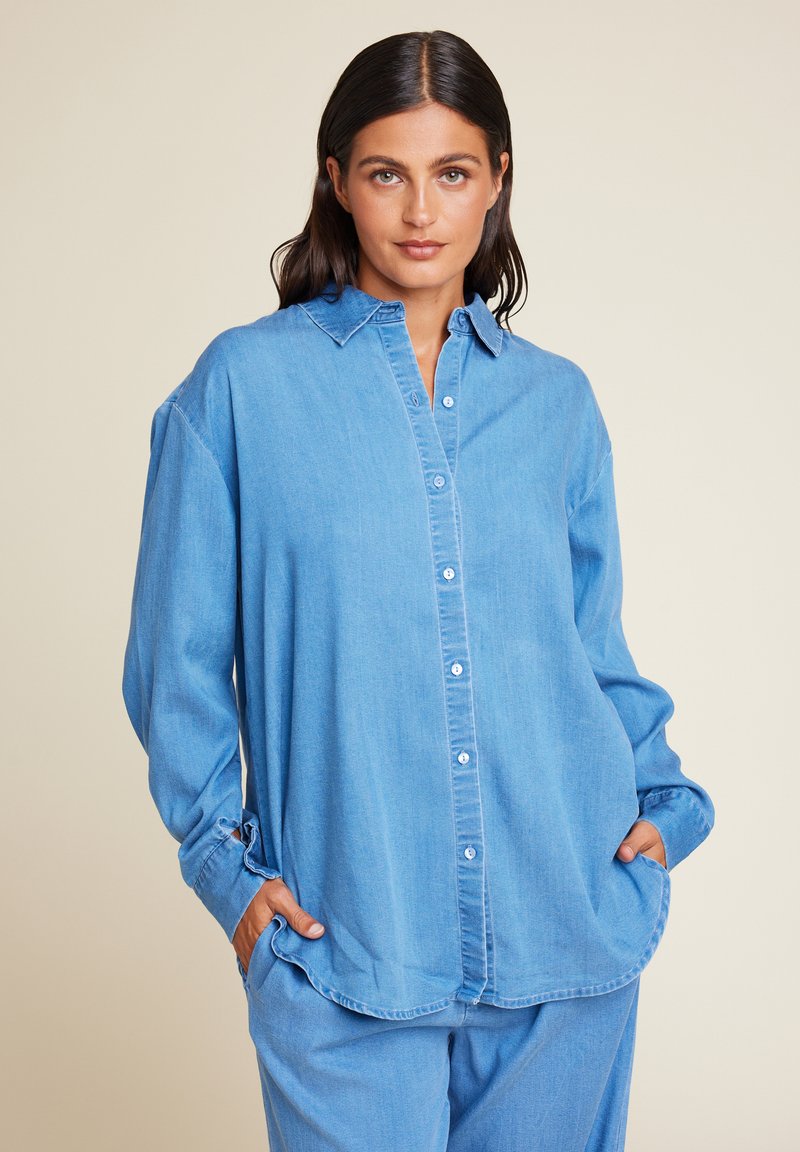 Lys blå denimskjorte med knapper foran, lange ermer og klassisk krage. Har lommer og en løs passform for komfort.