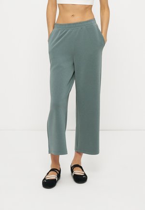Femme portant un pantalon de survêtement vert pâle, ample et court, avec une taille élastique, et des ballerines noires à brides, sur fond blanc.