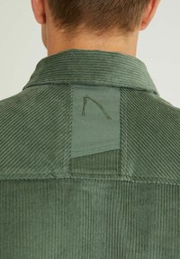 Vihreä corduroy-paita, jossa selkeä pystysuora ribbikuviointi, klassinen kaulus ja neliömäinen, takana oleva kirjailtu merkki.