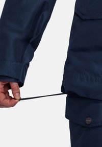 Marineblaue, wasserdichte Jacke mit einer Hand, die einen Kordelzug in der Taille anpasst. Glatte Textur mit einem Knopfdetail an der Seite.