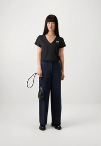 Kobieta z długimi prostymi czarnymi włosami, ubrana w czarną koszulkę z dekoltem w serek, granatowe szerokie spodnie, czarne buty i trzymająca małą czarną torebkę za pasek.