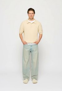 Beige ribbad pikétröja med korta ärmar, kombinerad med ljusblå jeans med avslappnad passform och beige slip-onskor. Enkel design, minimala detaljer.