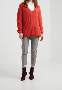 Červený pletený svetr s výstřihem do V a širokými rukávy, doplněný šedými kostkovanými tailored kalhotami a burgundskými kotníkovými botami.