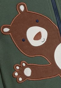 Brūnā lāča aplikācija uz zaļā ribotā auduma, ar baltām un zilo akcentiem, izšūtiem elementiem un rotaļīgu pēdas dizainu.