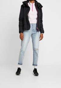 Schwarze, wasserdichte Jacke mit Kapuze, pinken Reißverschlüssen, getragen über einem hellrosa Pullover. Hellblaue, ausgebleichte Jeans und schwarze Sneaker runden das Outfit ab.