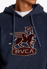 Sudadera con capucha de felpa en color navy, que presenta un logo bordado de grifo en burdeos, gris y blanco, con cordones ajustables y una textura suave.