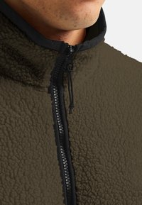 Brown fleece jacket with a textured surface, featuring a black zip closure and collar. The zipper includes a pull tab for easy fastening.