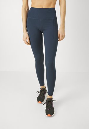 Hoch taillierte, navy-farbene Leggings aus dehnbarem Stoff mit glatter Textur und zwei Seitentaschen. Kombiniert mit schwarzen Sportschuhen.