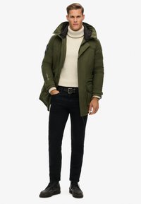 Olive zelena puhovka s kapuco, zadrgo in gumbom, z zadrgo zaprti žepi, črnimi detajli, nosi se čez smetanasto turtleneck majico in črne kavbojke.