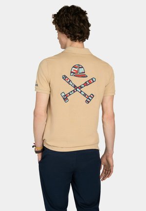 Hombre con camisa polo beige con diseño en la espalda de mallets de polo a rayas cruzados y casco, combinado con pantalones azul oscuro.