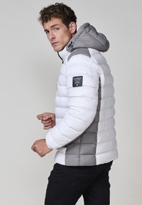 Giacca puffer bianca e grigia con cappuccio, caratterizzata da un design trapuntato, accenti sui pannelli laterali e una patch con logo sul braccio sinistro.