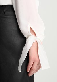 Blusa blanca de manga larga con tejido texturizado, que presenta una abertura y un detalle de lazo en los puños, combinada con una prenda negra debajo.