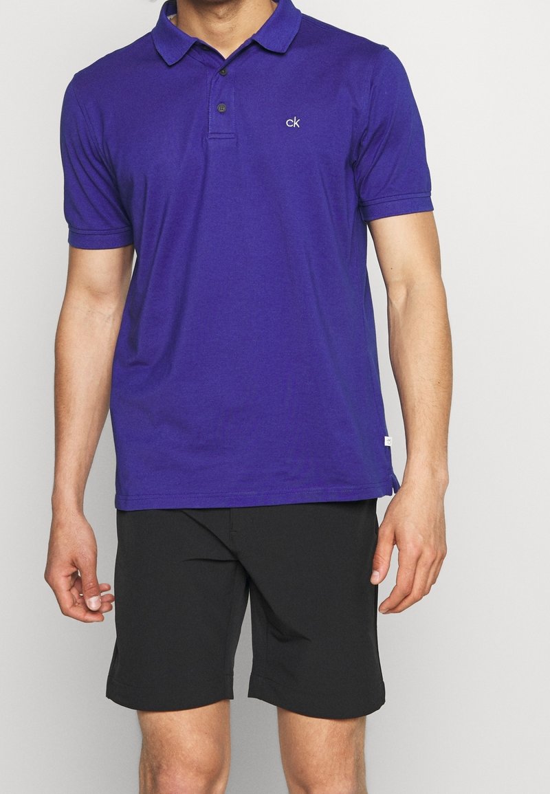Homme portant un polo à manches courtes violet avec le logo « ck » sur la poitrine et un short noir jusqu’aux genoux, debout devant un fond uni.