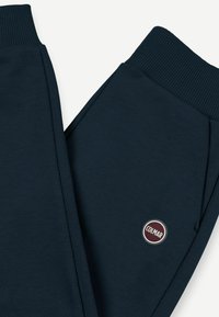 Tamsiai mėlynos treniruočių kelnės, pagamintos iš lygaus audinio, su raukiniais pavadėlyje ir apvaliu logotipu ant šlaunies.