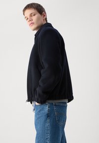 Navy blue bomber jacket with a zip front, ribbed cuffs, and hem. Paired with light blue jeans, featuring a straight-leg design and pockets.