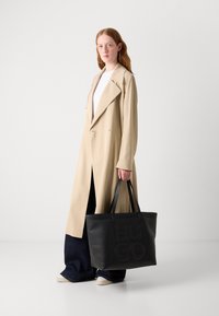 Schwarze Leder-Tasche mit eingeprägtem "HUGO"-Logo, ausgestattet mit zwei stabilen Griffen. Das Modell trägt einen beigen Trenchcoat über einem weißen Oberteil und dunklen Jeans.